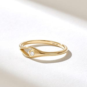 May include: A polished 14k gold ring featuring a smooth, tapered band that curves into an eye-shaped setting. A single round, clear gemstone is bezel-set within the center of the eye motif. The interior of the band is engraved with DIA 14K.