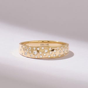 Könnte beinhalten: Ein goldener Bandring mit einem verstreuten Muster aus zahlreichen kleinen, runden, klaren Edelsteinen, die in die Außenfläche eingelassen sind. Die Innenseite des Rings ist deutlich mit dem Text DIA 14K graviert, was die Metallzusammensetzung angibt.