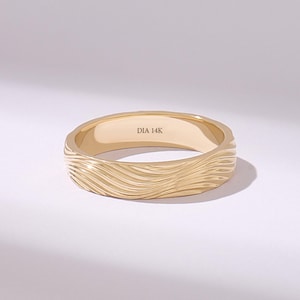 May include: A gold band ring featuring a textured exterior with flowing, wave-like horizontal grooves. The interior of the ring is smooth and clearly engraved with the text DIA 14K, indicating the metal composition of the jewelry piece.
