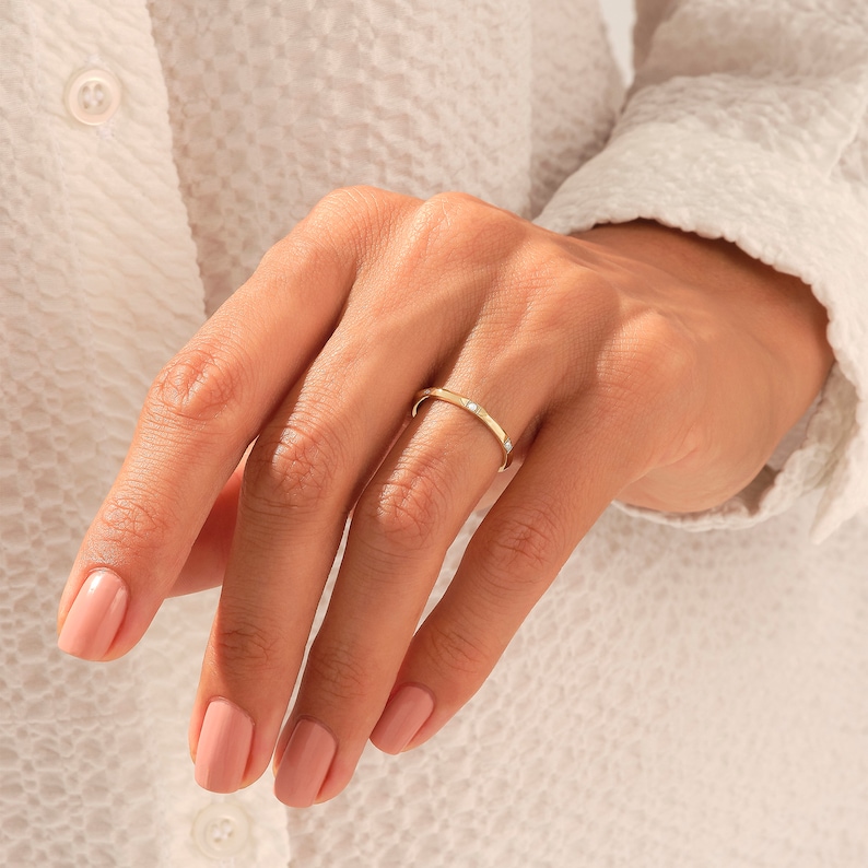 May include: A thin gold band ring featuring three small, evenly spaced, round, clear stones set flush into the metal surface. The ring has a polished finish and a simple, slender profile that encircles the finger, showcasing a clean design.