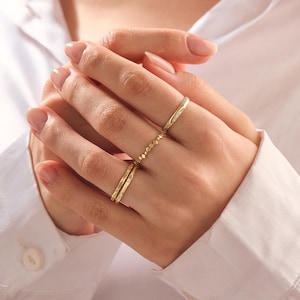 May include: A set of three gold-toned rings worn on the fingers. The top ring features a thin band with small embedded stones. The middle ring has a textured, beaded design. The bottom ring is a smooth, thin band with a single small stone accent.