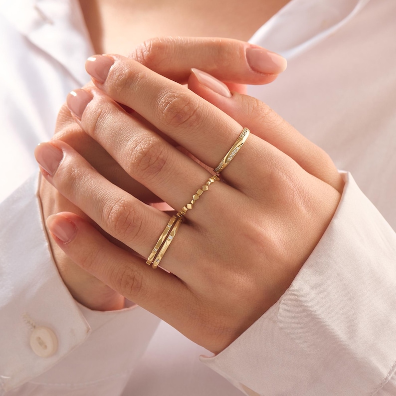 May include: A set of three gold-toned rings worn on the fingers. The top ring features a thin band with small embedded stones. The middle ring has a textured, beaded design. The bottom ring is a smooth, thin band with a single small stone accent.