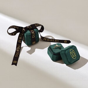 以下が含まれることがあります： 「DIA FINE JEWELRY」と金色の文字が書かれた緑色のベルベット製のリングボックスが2つ。1つのボックスが開いており、透明な石がついた金のリングが入っています。