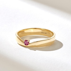 Op de afbeelding: Een gouden ring met een kleine, ronde, rode edelsteen. De ring heeft een glad, gepolijst oppervlak en een uniek ontwerp. De tekst "DIA 14K" is aan de binnenkant van de ring gestempeld. De ring staat tegen een witte achtergrond.