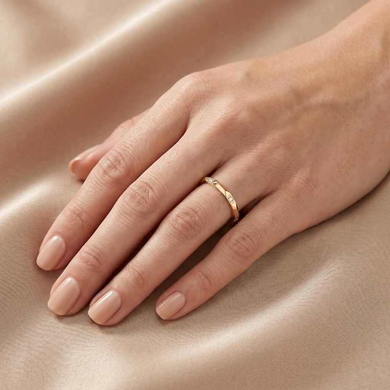 May include: A gold band ring featuring a faceted geometric design with small, clear, square-cut stones embedded at intervals along the outer surface. The ring has a polished metallic finish and sits on a finger, showcasing its angular, modern profile.