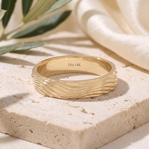 May include: A gold band ring featuring a textured, wavy surface pattern on the exterior. The interior of the ring is smooth and clearly engraved with the text DIA 14K, indicating the metal composition and material quality of the jewelry piece.