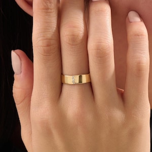 Puede incluir: Un anillo de banda de oro pulido que presenta una fila vertical de tres peque&ntilde;as piedras redondas y transparentes engastadas en el centro de la superficie met&aacute;lica. El anillo tiene un acabado liso y plano con un ancho constante.