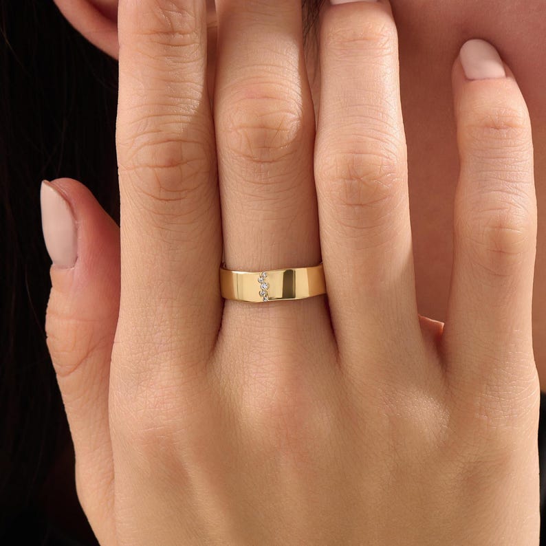 Puede incluir: Un anillo de banda de oro pulido que presenta una fila vertical de tres peque&ntilde;as piedras redondas y transparentes engastadas en el centro de la superficie met&aacute;lica. El anillo tiene un acabado liso y plano con un ancho constante.
