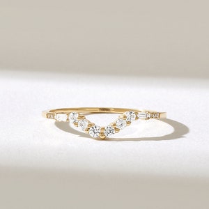May include: A gold ring with a curved design, set with multiple round and baguette-cut diamonds. The ring has a delicate band and is displayed on a white surface. The text "DIA 14K" is visible on the ring.