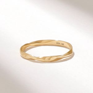 May include: A gold ring with a twisted, undulating band design. The interior surface of the band features the engraved text DIA 14K. The metal has a polished, reflective finish that catches the light, creating subtle highlights along the curves.