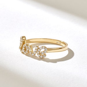 May include: A gold ring spelling out the word "love" in cursive script. The letters are encrusted with small, sparkling gemstones. The ring band is a simple, polished gold. The inscription "DIA 14K" is visible on the band.