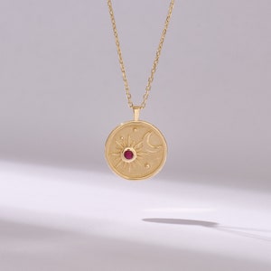 Op de afbeelding: Gouden ketting met een ronde hanger met een zon- en maandesign met een rode edelsteen in het midden.