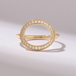 以下が含まれることがあります： 大きな楕円形の開口部を持つゴールドリング。小さなダイヤモンドが縁に沿って埋め込まれています。細いバンドには「DIA 14K」の刻印があります。白い背景に表示されています。モダンでエレガントなデザイン。