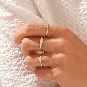 May include: Three gold rings worn on fingers. The top ring is a thin band with two small embedded stones. The middle ring features an elongated oval plate with a single centered stone. The bottom ring has a cluster of three small stones.