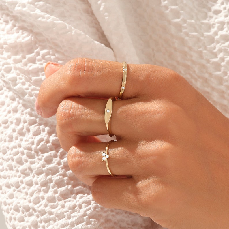 May include: Three gold rings worn on fingers. The top ring is a thin band with two small embedded stones. The middle ring features an elongated oval plate with a single centered stone. The bottom ring has a cluster of three small stones.