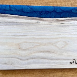 May include: A wooden cutting board with a blue epoxy resin edge. The board has a natural wood grain pattern and the text "FAT CAT" is engraved on the bottom right corner.