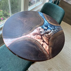 May include: Round wooden table with a dark brown finish and a unique epoxy resin design. The resin flows through the wood in shades of blue, copper, and white. The table is approximately 24 inches in diameter.