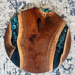 May include: A circular table top made from a natural wood slab with a prominent grain pattern and a dark brown finish. Two irregular channels filled with a swirling, metallic teal and dark green epoxy resin run through the wood surface.