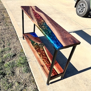 May include: A long, wooden console table with a black metal frame. The table top features a colorful epoxy river design with blue, green, gold, and red hues.