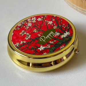 Pode incluir: Caixa de pílulas dourada com um design floral vermelho e branco e o nome "Poppy" escrito em letras douradas.