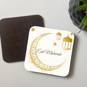 Op de afbeelding: Vierkante onderzetter met een bruine achterkant en een witte bovenkant met een gouden halve maan, lantaarns en sterren. De woorden "Eid Mubarak" zijn in elegant schrift geschreven. De onderzetter is ontworpen voor dranken.