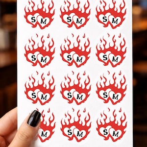 May include: Sheet of stickers featuring a red flame heart design with the letters "S" and "M" inside each heart. The stickers are on a white background and held by a hand.