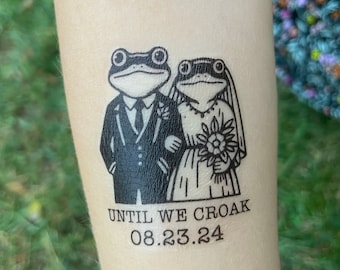 May include: A black and white temporary tattoo of two frogs dressed in wedding attire. The frog groom is wearing a suit and tie, and the frog bride is wearing a dress and veil. The text "Until We Croak 08.23.24" is below the frogs.