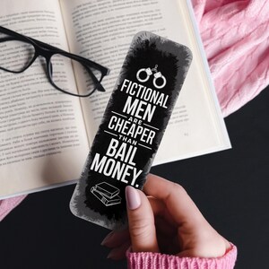 May include: A black and white bookmark with the text "FICTIONAL MEN ARE CHEAPER THAN BAIL MONEY." The bookmark features a pair of handcuffs and a stack of books. The bookmark is held in front of an open book and glasses.