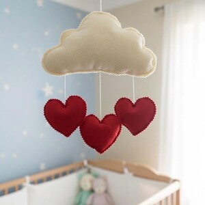 May include: A baby mobile featuring a cream-colored cloud with a scalloped edge, suspended above a crib. Three red hearts with scalloped edges hang below the cloud. The nursery has a light blue wall with white stars.