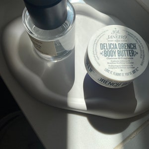 Peut inclure: Un plateau en c&eacute;ramique blanche avec une bouteille de parfum en verre transparent et un pot de beurre corporel blanc. La bouteille de parfum a un bouchon noir et le pot de beurre corporel a une &eacute;tiquette noire et blanche avec le texte "Sol de Janeiro Delicia Drench Body Butter" et "Cheirosa 59 Fragrance".