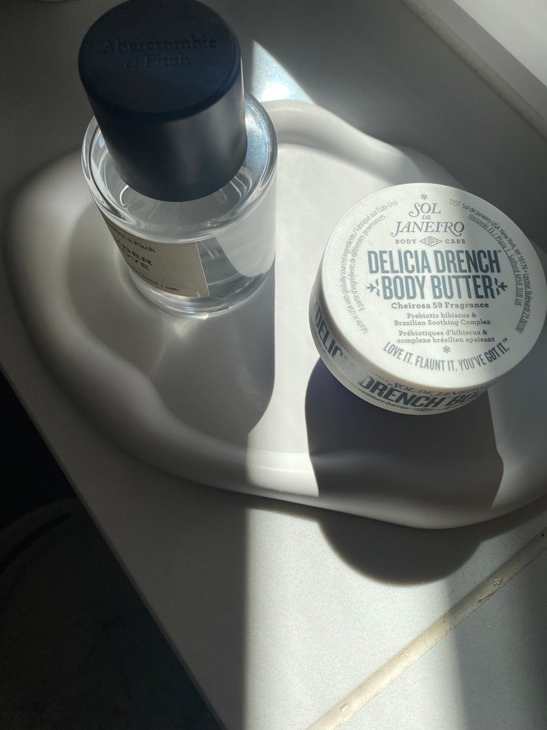 Peut inclure: Un plateau en c&eacute;ramique blanche avec une bouteille de parfum en verre transparent et un pot de beurre corporel blanc. La bouteille de parfum a un bouchon noir et le pot de beurre corporel a une &eacute;tiquette noire et blanche avec le texte "Sol de Janeiro Delicia Drench Body Butter" et "Cheirosa 59 Fragrance".