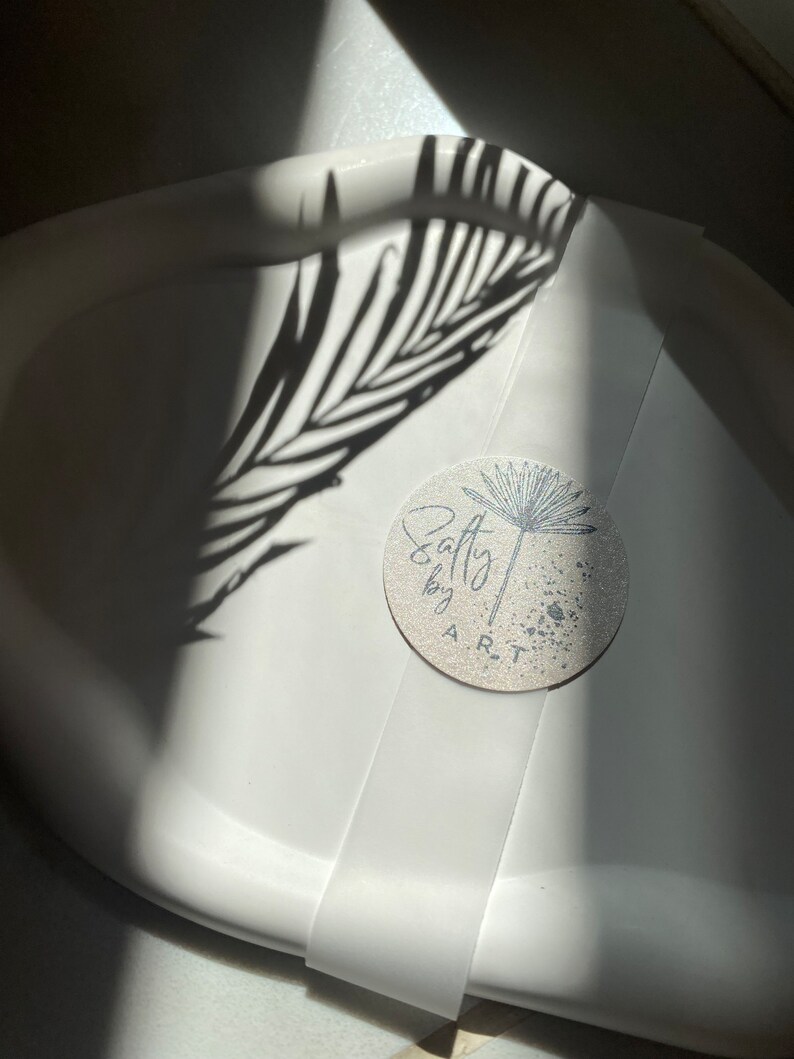 Peut inclure: Un plateau de service en c&eacute;ramique blanche avec un autocollant rond qui dit "Salty by ART" dans une police d'&eacute;criture manuscrite. L'autocollant a un motif de pissenlit noir et blanc et un fond mouchet&eacute;.