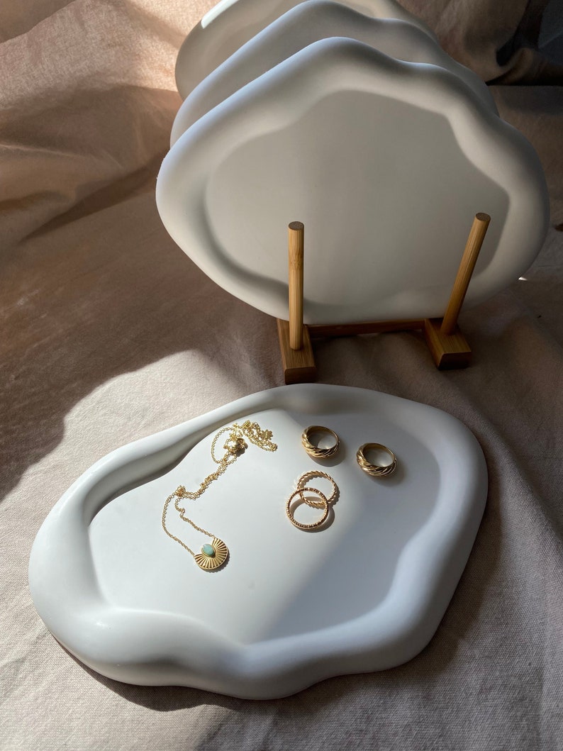 Peut inclure: Plateau en c&eacute;ramique blanche avec un bord ondul&eacute;, pr&eacute;sentant un collier en or avec un pendentif en forme de soleil et trois anneaux en or. Le plateau est sur un fond en tissu blanc.