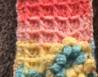 May include: A crocheted wrist warmer with a flower design. The wrist warmer is made of pink, yellow, and blue yarn.