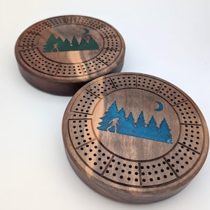 May include: Two wooden cribbage boards featuring a forest scene design. The boards are brown with a dark wood grain. The forest scene is green and blue with a crescent moon.
