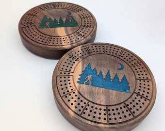 May include: Two wooden cribbage boards with a forest scene design. The boards are brown with a dark wood grain. The forest scene is green and blue with a crescent moon.