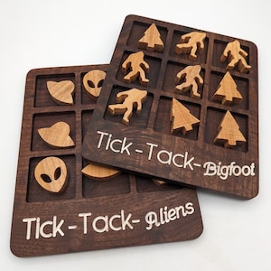 May include: Two wooden tic-tac-toe boards with different themes. One board features aliens, UFOs, and hearts, and the other features Bigfoot, trees, and footprints. The boards are made of dark wood with white text.
