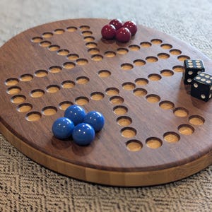 May include: A wooden board game with a dark brown playing surface and a light brown rim. The game includes red and blue marbles, and two black dice with white dots. The board has a series of holes for the marbles.