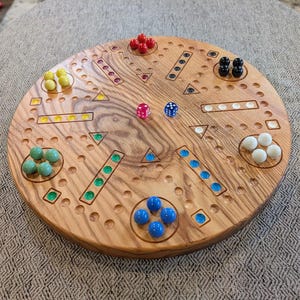 Puede incluir: Un tablero de juego de madera circular con una variedad de canicas y dados de colores. El tablero tiene un patrón de líneas y círculos, y las canicas están dispuestas en grupos de tres.