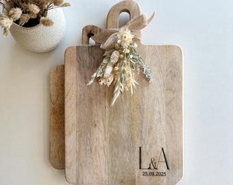 Puede incluir: Dos tablas de cortar rectangulares de madera clara con asas, decoradas con un arreglo de flores secas atado con una cinta de arpillera. La tabla frontal tiene grabadas las letras L & A y la fecha 25.09.2025 en una fuente negra.