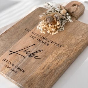 Könnte beinhalten: Ein rechteckiges Holzschneidebrett mit Griff, verziert mit einem Trockenblumenarrangement und einer Juteschleife. Das Brett ist mit dem Text DIE GEHEIME ZUTAT IST IMMER Liebe sowie den Namen JULIA & SIMON und dem Datum 12.07.2026 graviert.