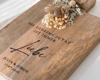 May include: A rectangular wooden cutting board with a handle, featuring a dried floral arrangement tied with a burlap ribbon. The board is engraved with the German text DIE GEHEIME ZUTAT IST IMMER Liebe, followed by the names JULIA & SIMON and 12.07.2026.