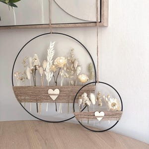 Könnte beinhalten: Zwei kreisförmige Wandbehänge mit Trockenblumen. Jeder hat einen schwarzen Metallring, eine mit Bindfaden umwickelte Basis und ein Holzherz. Der größere hat eine Vielzahl von beigen und weißen Blumen. Der kleinere hat ein ähnliches Design mit weniger Blumen.