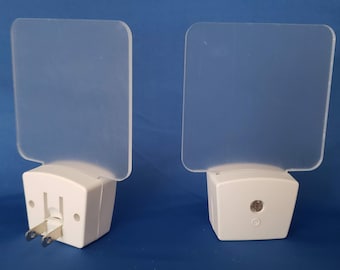 Könnte beinhalten: Zwei weiße Steckdosen-Nachtlichter mit mattierten quadratischen Lichtabdeckungen. Die Lichter haben einen kleinen Sensor an der Vorderseite und einen Schalter an der Seite.