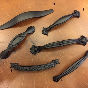 May include: A collection of six black cabinet pulls and handles in various designs. Styles include curved, ornate, and ribbon-shaped options. The hardware is displayed on a wooden surface.