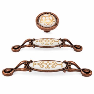 May include: A set of three cabinet hardware pieces including one round knob and two elongated drawer pulls. Each piece features a white ceramic centre with a gold filigree pattern, encased in an ornate, dark copper-toned metal frame.