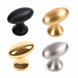 May include: A set of four oval-shaped cabinet knobs featuring different finishes. One knob is matte black, one is brushed gold, one is polished gold, and one is brushed silver. Each knob has a rounded base and a smooth, elongated top surface.