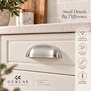 May include: A silver-toned metal cup pull handle mounted on a cream-colored cabinet drawer. The image includes text stating Small Details. Big Difference. and GÜRGAY HOME HARDWARE, along with icons for premium zinc alloy, multiple sizes, and screws included.