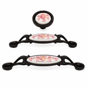 May include: A set of three cabinet hardware pieces including one round knob and two elongated drawer pulls. Each piece features a matte black metal frame with a central white ceramic insert decorated with a pink and gold floral pattern.