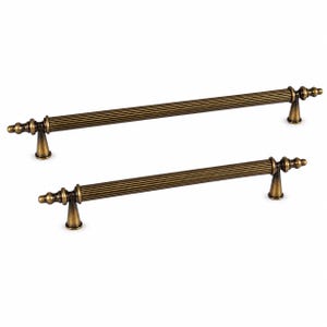 May include: Two antique brass cabinet pulls featuring a long, cylindrical, fluted bar design. Each handle is supported by two conical pedestals and finished with decorative finials at both ends, showcasing a metallic, weathered gold-toned finish.